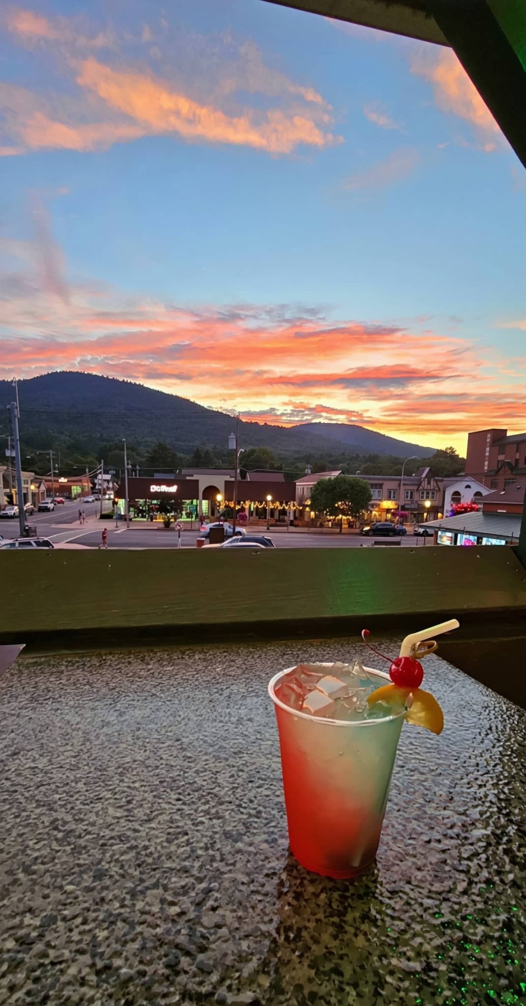 A cherry-garnished cocktail on the deck railing with a colorful sunset over Lake George village