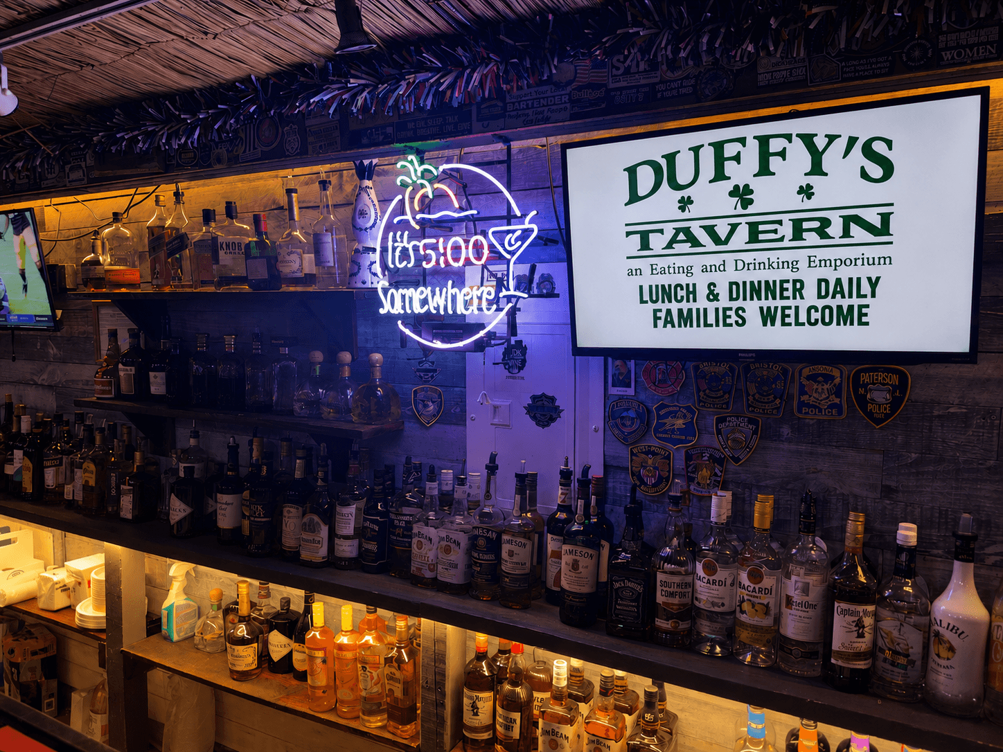 Backlit liquor shelves at Duffy's bar, with a neon sign and a screen welcoming families in for lunch and dinner