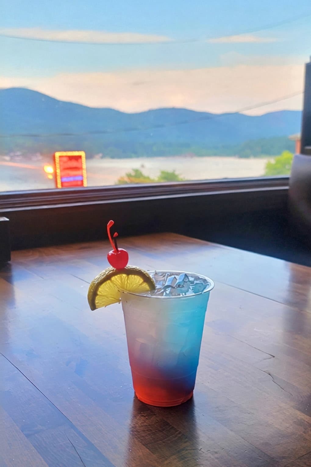 A red-and-blue layered cocktail with lemon and cherry garnish on a table overlooking Lake George at dusk