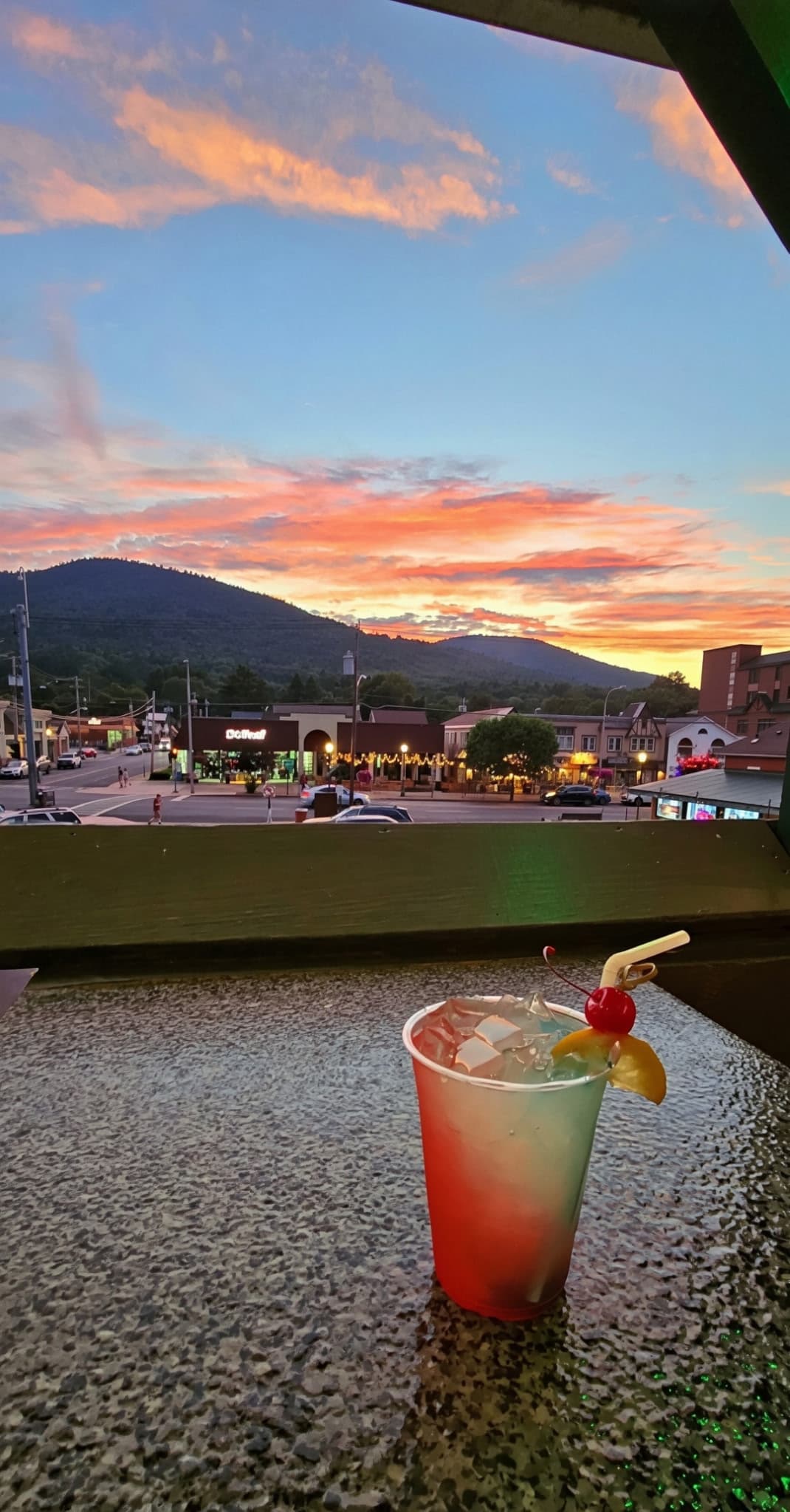A cherry-garnished cocktail on the deck railing with a colorful sunset over Lake George village